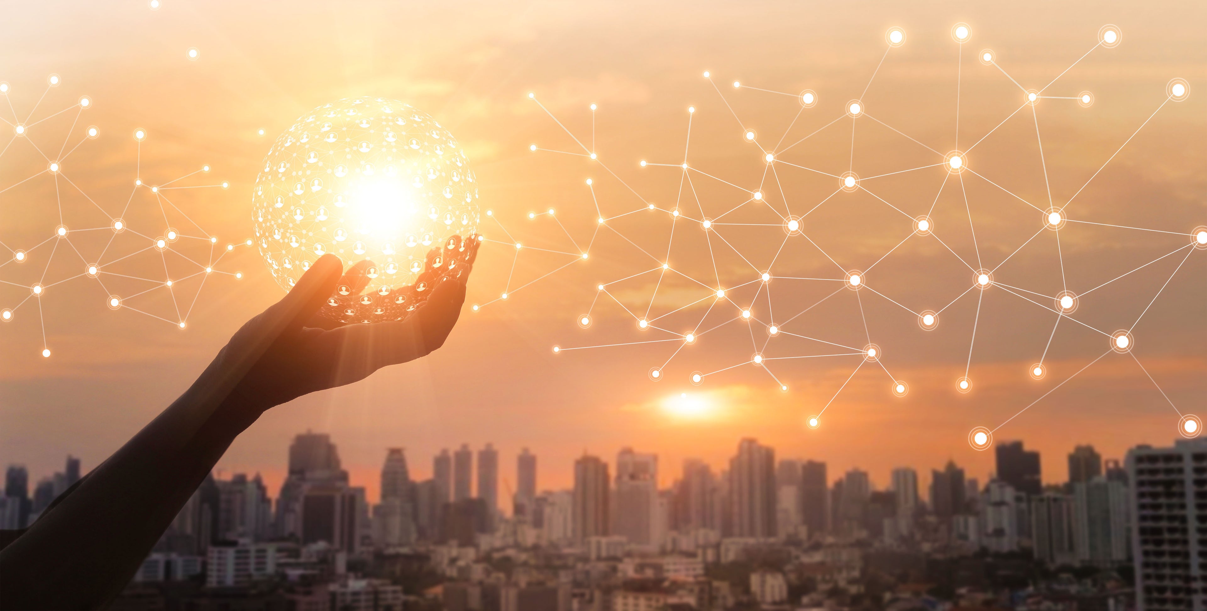 Hand holding a glowing sphere with network connections over a cityscape at sunset.