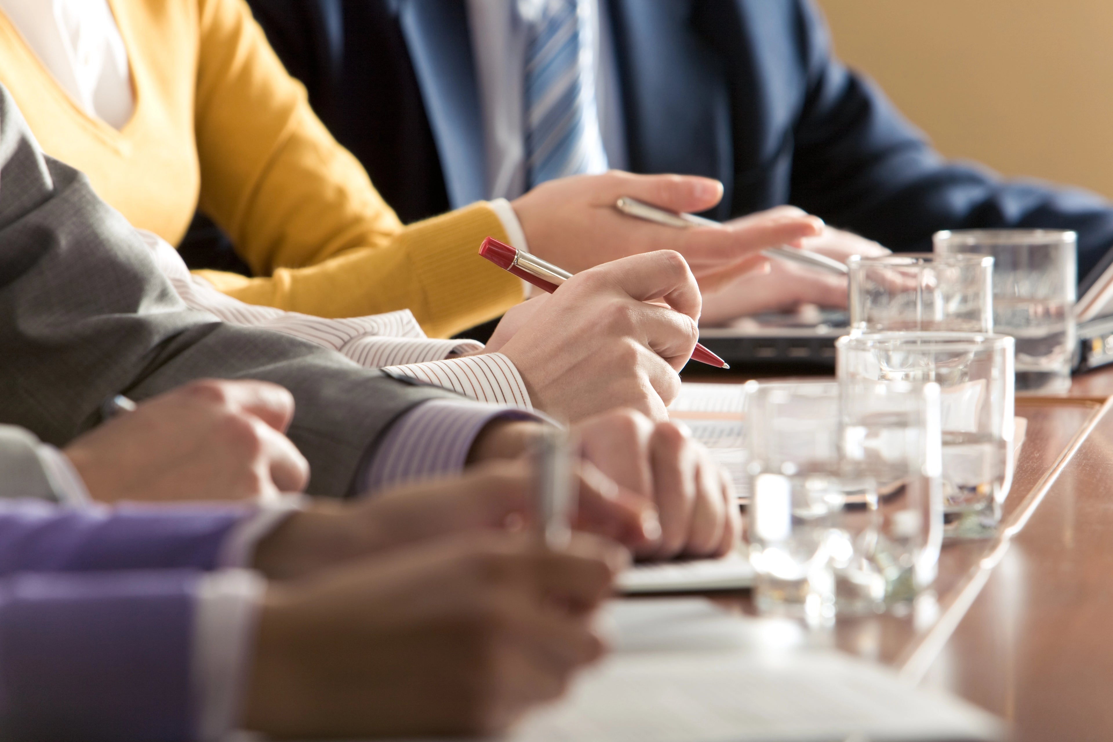 People sitting at a table with hands and pens visible, likely in a meeting or discussion setting.